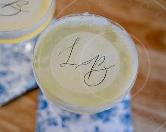 May include: Overhead view of two cocktails in stemmed glasses. The drinks have a pale yellow hue and are topped with a foamy layer. Each glass has a circular sticker with the cursive letters "LB". The glasses rest on blue and white patterned napkins.