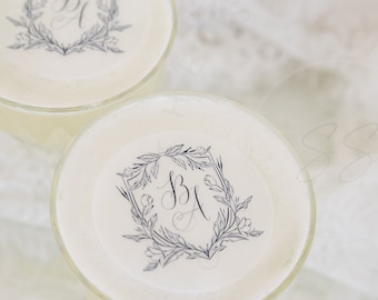 May include: Two clear glasses filled with a creamy beige liquid, each topped with a circular white garnish featuring a dark blue printed design of the letters BA enclosed within a rectangular frame of delicate botanical leaves and flowers.