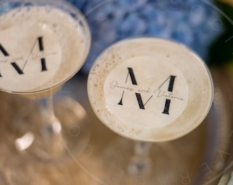 May include: Two martini glasses filled with a beige, foamy cocktail. Each drink features a circular edible topper printed with a large black letter M, the text James and Victoria, and the Roman numeral IV in a serif font style.