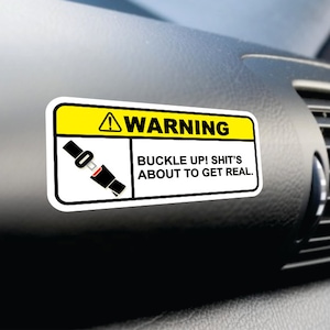 Könnte beinhalten: Ein gelber und weißer Warnhinweis mit einer schwarzen und roten Illustration eines Sicherheitsgurts. Der Aufkleber lautet "WARNING BUCKLE UP! SHIT'S ABOUT TO GET REAL."