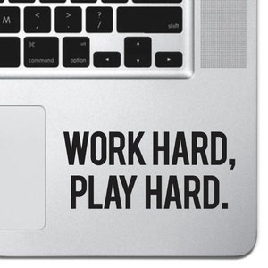 Puede incluir: Un portátil plateado con una pegatina negra que dice "TRABAJA DURO, JUEGA DURO."