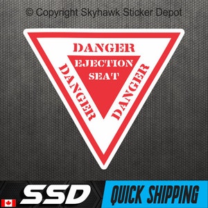 Puede incluir: Una pegatina triangular blanca y roja con las palabras "DANGER EJECTION SEAT" en negrita, fuente sans-serif. La palabra "DANGER" se repite a lo largo de los lados del triángulo. La pegatina tiene un fondo negro.