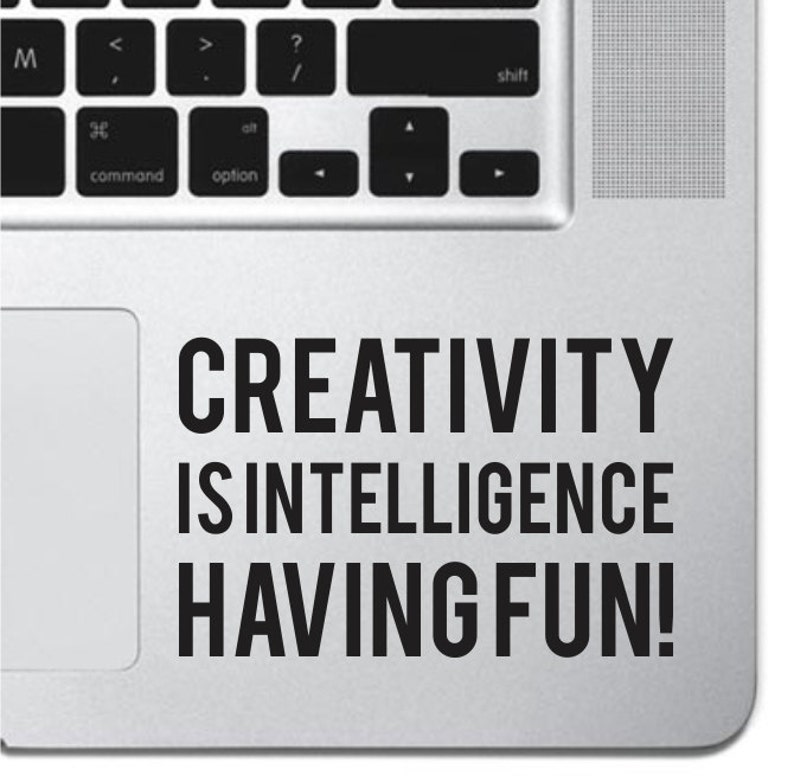May include: A silver laptop with a black sticker that reads "CREATIVITY IS INTELLIGENCE HAVING FUN!"