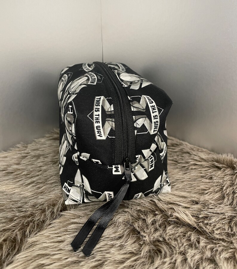 May include: Black cosmetic bag with a repeating pattern of white and gray images and the text "THIS IS THE WAY". The bag has a black zipper and a black ribbon pull.