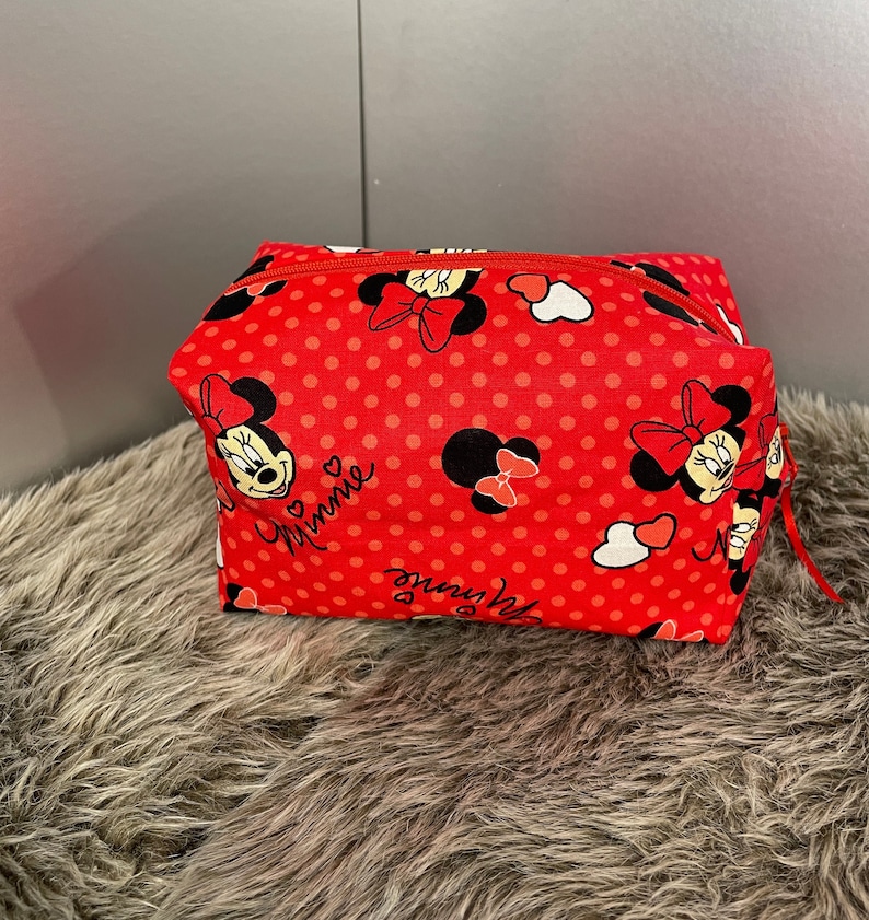 Op de afbeelding: Rode toilettas met rits, stippenpatroon en afbeeldingen van Minnie Mouse. De tas heeft een wit hart en de tekst "Minnie" erop.