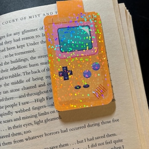 May include: A glittery orange and pink bookmark shaped like a handheld video game console. The bookmark has a screen, buttons, and a directional pad.