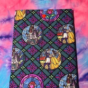 Könnte beinhalten: Ein rechteckiger Stoffartikel mit einem Buntglasdesign, das ein Schloss, eine Rose und ein tanzendes Paar zeigt. Das Design ist vor einem teal-farbenen Hintergrund mit einem geometrischen Muster angeordnet, und der Hintergrund ist ein Tie-Dye aus Pink und Blau.