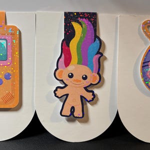 Könnte beinhalten: Drei farbenfrohe Lesezeichen mit einer Retro-Handheld-Spielkonsole, einem Regenbogen-Troll und einem lila und rosa Tamagotchi-ähnlichen Spielzeug. Jedes Lesezeichen ist mit Glitzer verziert.