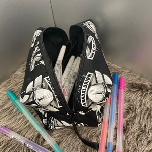 May include: Black pencil case with a zipper, featuring a repeating pattern of The Mandalorian helmet and text. The case is open, revealing white markers and several colorful gel pens in purple, teal, pink, and blue.