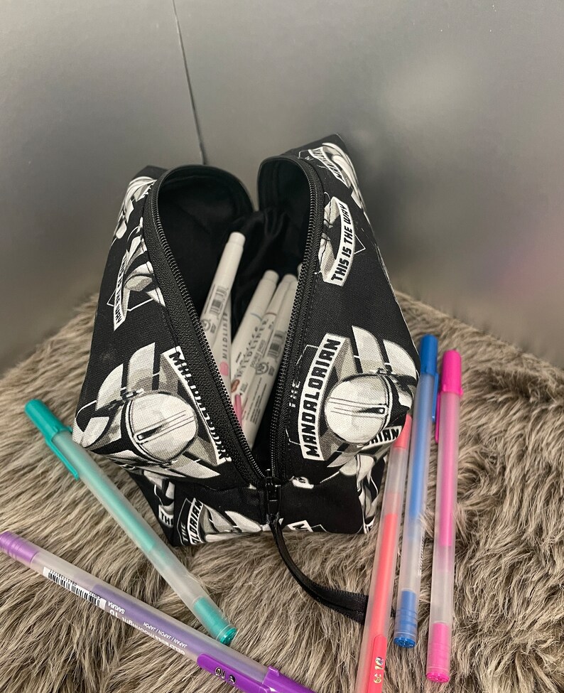 May include: Black pencil case with a zipper, featuring a repeating pattern of The Mandalorian helmet and text. The case is open, revealing white markers and several colorful gel pens in purple, teal, pink, and blue.
