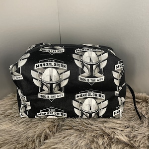 May include: Black cosmetic bag featuring a repeating pattern of The Mandalorian helmet and text "This is the Way". The bag has a black zipper and a small black loop on the side. The bag is sitting on a brown, furry surface.