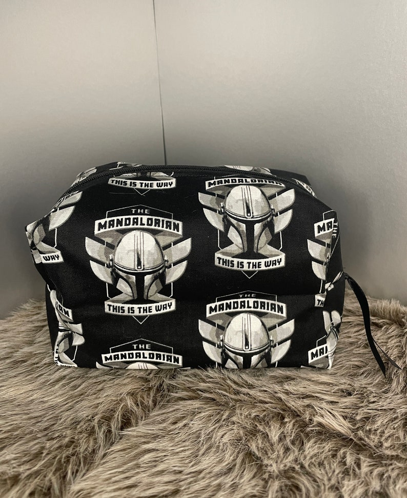 May include: Black cosmetic bag featuring a repeating pattern of The Mandalorian helmet and text "This is the Way". The bag has a black zipper and a small black loop on the side. The bag is sitting on a brown, furry surface.