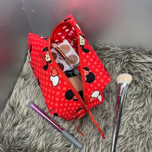 Op de afbeelding: Rode toilettas met witte stippen, een ritssluiting en een Minnie Mouse-ontwerp. De tas is open en bevat make-upkwasten en een make-uppalet.