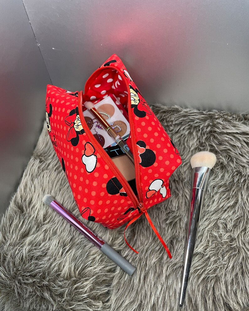 Op de afbeelding: Rode toilettas met witte stippen, een ritssluiting en een Minnie Mouse-ontwerp. De tas is open en bevat make-upkwasten en een make-uppalet.