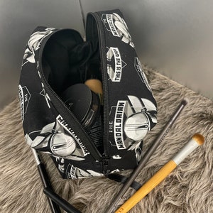 May include: Black cosmetic bag with a zipper, featuring a repeating pattern of a Mandalorian helmet and the text "THIS IS THE WAY" and "THE MANDALORIAN". Several makeup brushes and compacts are visible.