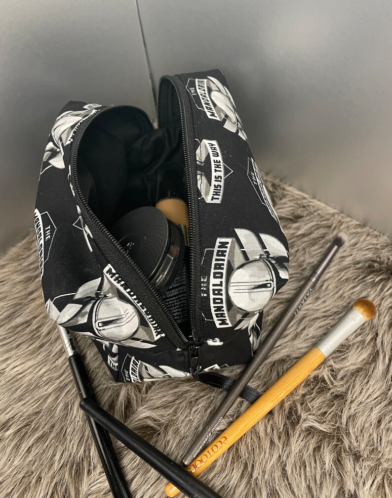 May include: Black cosmetic bag with a zipper, featuring a repeating pattern of a Mandalorian helmet and the text "THIS IS THE WAY" and "THE MANDALORIAN". Several makeup brushes and compacts are visible.