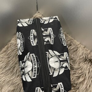 May include: Black rectangular zippered pouch featuring a repeating pattern of The Mandalorian helmet and the phrase "THIS IS THE WAY" in white. The pouch has a black zipper and is likely made of fabric.