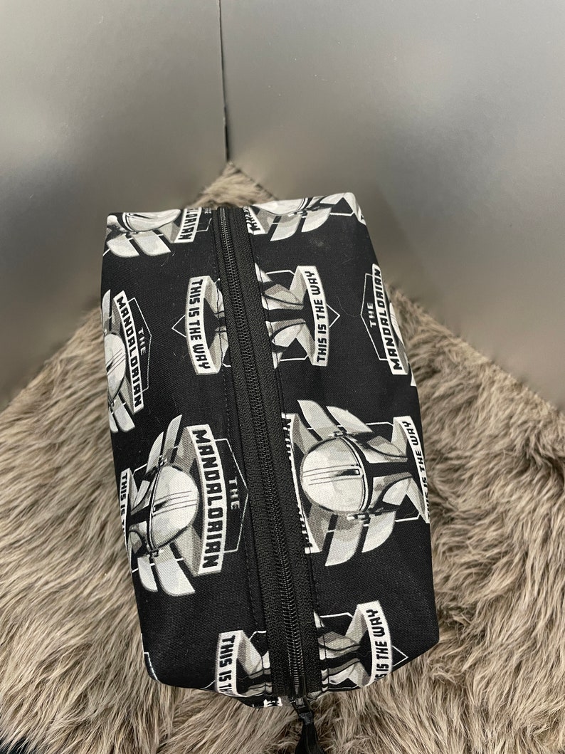 May include: Black rectangular zippered pouch featuring a repeating pattern of The Mandalorian helmet and the phrase "THIS IS THE WAY" in white. The pouch has a black zipper and is likely made of fabric.