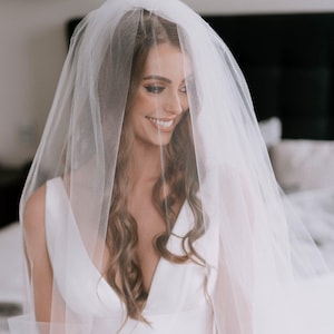 May include: A bride wearing a white sleeveless wedding dress with a long white veil. She is sitting on a bed and looking down.
