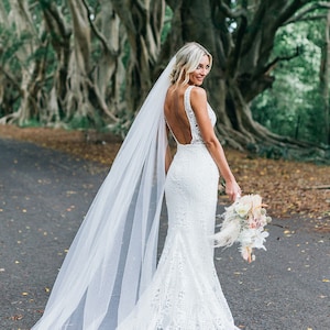 Blonde bride stands on a tree lined country lane looking over her shoulder. Brides veil is a 108 inch cathedral length that flows a bit past her dress train. The veil is sheer to show off the back details of her dress, which is our specialty style.