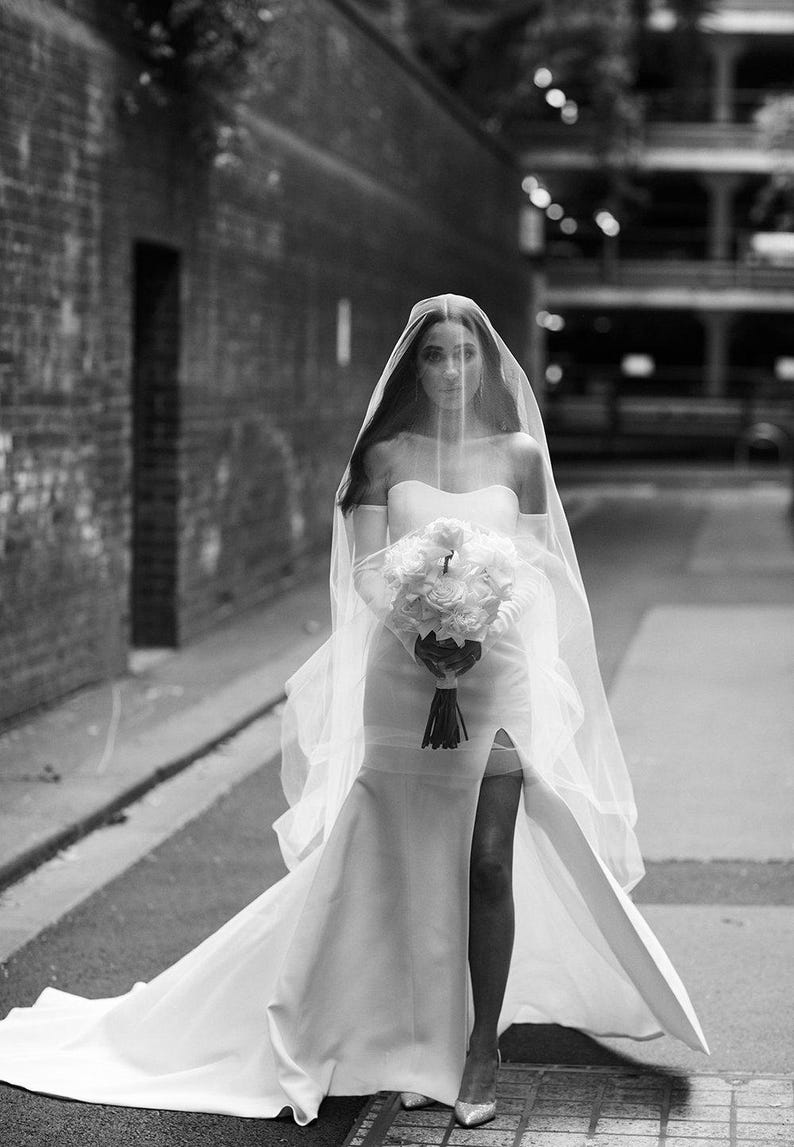 May include: A bride wearing a white strapless wedding dress with a long train and a veil. She is holding a bouquet of white flowers.