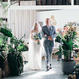 K&ouml;nnte beinhalten: Eine Braut in einem wei&szlig;en Spitzen-Hochzeitskleid geht mit ihrem Vater den Gang entlang. Sie tr&auml;gt einen Strau&szlig; aus wei&szlig;en und blauen Blumen. Die Zeremonie findet in einem modernen, minimalistischen Raum mit viel Gr&uuml;n statt.