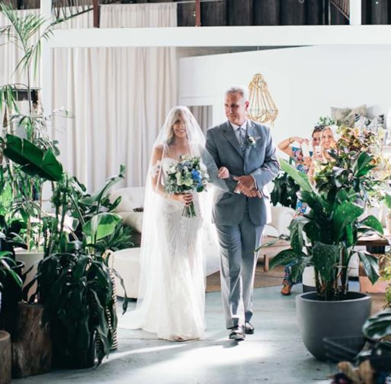 K&ouml;nnte beinhalten: Eine Braut in einem wei&szlig;en Spitzen-Hochzeitskleid geht mit ihrem Vater den Gang entlang. Sie tr&auml;gt einen Strau&szlig; aus wei&szlig;en und blauen Blumen. Die Zeremonie findet in einem modernen, minimalistischen Raum mit viel Gr&uuml;n statt.