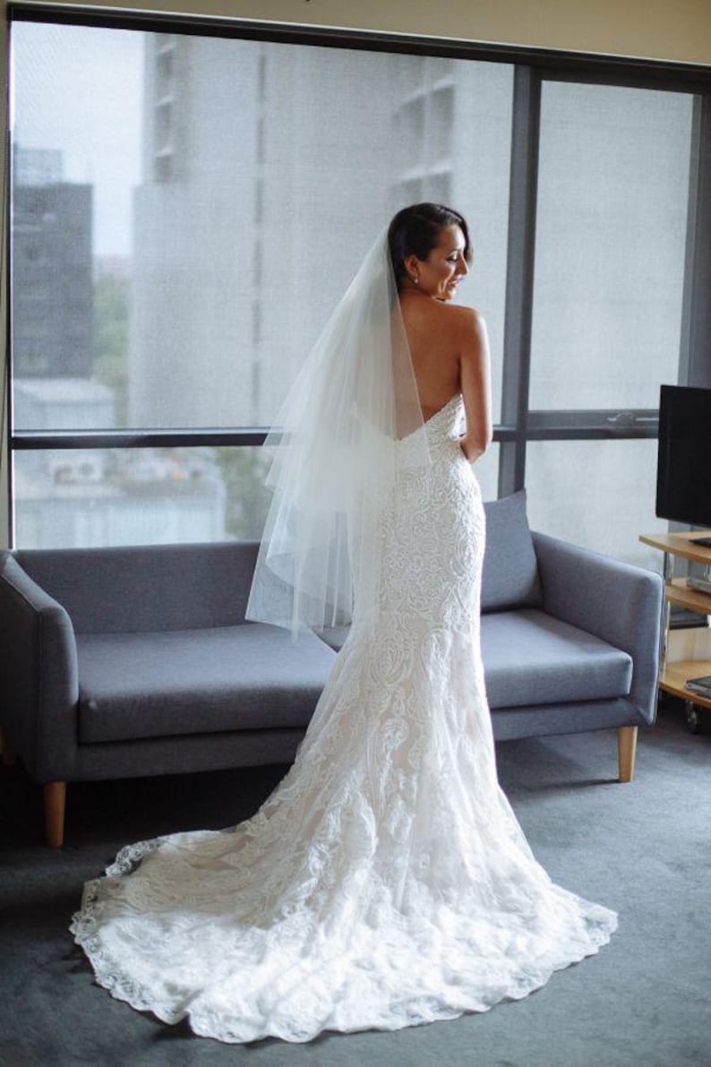 K&ouml;nnte beinhalten: Eine Braut in einem wei&szlig;en Spitzen-Hochzeitskleid mit langem Schleier und Schleppe. Sie steht vor einem Fenster mit einem Sofa hinter ihr.