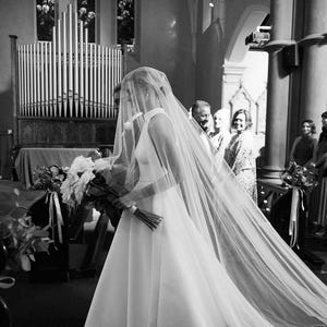 K&ouml;nnte beinhalten: Eine Braut in einem wei&szlig;en Hochzeitskleid mit langem Schleier geht den Gang einer Kirche entlang. Sie tr&auml;gt einen Strau&szlig; wei&szlig;er Blumen.