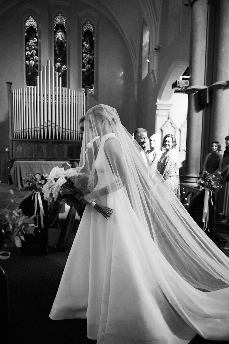 K&ouml;nnte beinhalten: Eine Braut in einem wei&szlig;en Hochzeitskleid mit langem Schleier geht den Gang einer Kirche entlang. Sie tr&auml;gt einen Strau&szlig; wei&szlig;er Blumen.