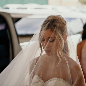 K&ouml;nnte beinhalten: Eine Braut in einem wei&szlig;en Hochzeitskleid und Schleier, die mit geschlossenen Augen nach unten schaut. Sie steht vor einem Auto, hinter ihr steht eine Brautjungfer.