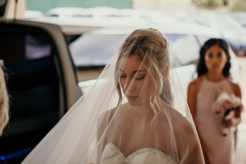 K&ouml;nnte beinhalten: Eine Braut in einem wei&szlig;en Hochzeitskleid und Schleier, die mit geschlossenen Augen nach unten schaut. Sie steht vor einem Auto, hinter ihr steht eine Brautjungfer.