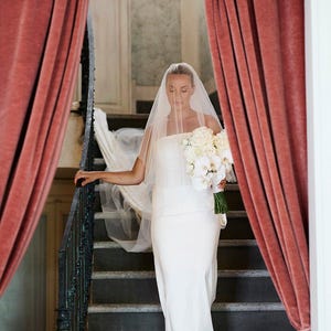 K&ouml;nnte beinhalten: Eine Braut in einem wei&szlig;en Hochzeitskleid und Schleier geht eine Treppe hinunter mit einem Strau&szlig; wei&szlig;er Blumen. Die Treppe wird von roten Samtvorh&auml;ngen eingerahmt.