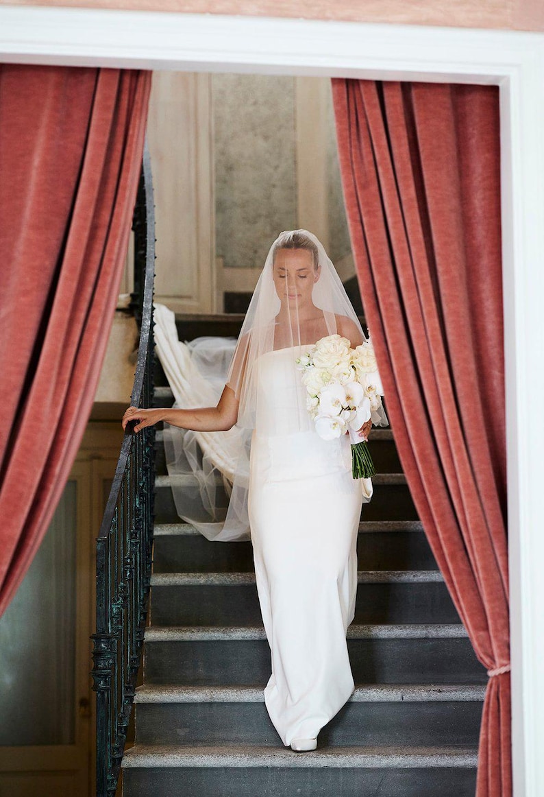 K&ouml;nnte beinhalten: Eine Braut in einem wei&szlig;en Hochzeitskleid und Schleier geht eine Treppe hinunter mit einem Strau&szlig; wei&szlig;er Blumen. Die Treppe wird von roten Samtvorh&auml;ngen eingerahmt.