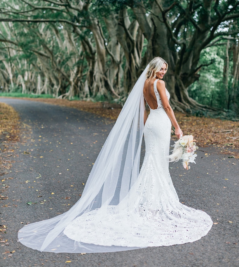 Blonde bride stands on a tree lined country lane looking over her shoulder. Brides veil is a 108 inch cathedral length that flows a bit past her dress train. The veil is sheer to show off the back details of her dress, which is our specialty style.