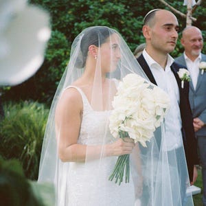 K&ouml;nnte beinhalten: Eine Braut in einem wei&szlig;en Hochzeitskleid und Schleier geht den Gang entlang mit einem Strau&szlig; wei&szlig;er Rosen. Der Br&auml;utigam steht hinter ihr und l&auml;chelt.