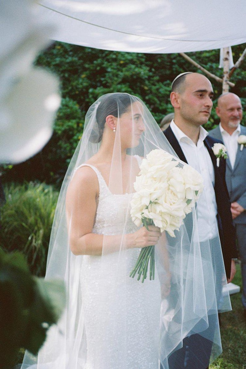K&ouml;nnte beinhalten: Eine Braut in einem wei&szlig;en Hochzeitskleid und Schleier geht den Gang entlang mit einem Strau&szlig; wei&szlig;er Rosen. Der Br&auml;utigam steht hinter ihr und l&auml;chelt.