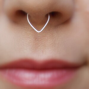 May include: A silver heart-shaped nose ring piercing. The ring is made of thin wire and is open on one side.