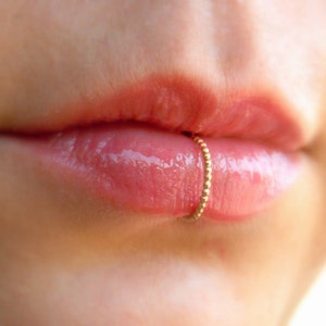 Può includere: Primo piano di labbra con un piercing dorato. L'anello è un piccolo cerchio perlato e le labbra sono lucide e rosa. La pelle intorno alle labbra è liscia e chiara.