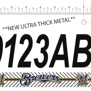 May include: A white metal license plate with black text that reads "**NEW ULTRA THICK METAL**" and "0123AB". The plate has a blue and gold striped design with a "M" logo on each side. The plate is 2.3 inches tall.