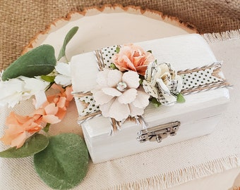 Puede incluir: Caja de anillo de madera blanca decorada con un diseño floral. La caja tiene una tapa con bisagras y un cierre de pestillo. Las flores están hechas de papel y tela y son en tonos de melocotón, blanco y verde.