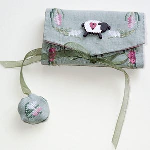 May include: A green fabric needle case with a ribbon closure and a wooden sheep button. The case is decorated with a floral design and has a small matching pincushion.