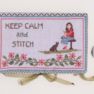 May include: A blue and white needlepoint pillow with a green ribbon. The pillow features a girl stitching with a cat beside her. The text on the pillow reads "Keep Calm and Stitch".