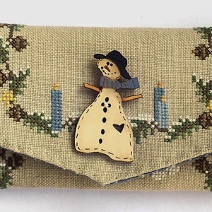 May include: A beige fabric pouch with a cross-stitch design of a snowman with a black hat and blue scarf, standing in front of two blue candles. The pouch is decorated with a green and brown leafy border.