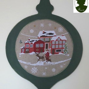Puede incluir: Un adorno de madera verde con forma de bola de Navidad con fondo blanco y un diseño de punto de cruz de un pueblo de invierno con Papá Noel y renos. El adorno tiene un lazo en la parte superior para colgar.