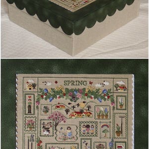 May include: A green and beige box with a scalloped edge. The top of the box features a cross-stitch design of a spring scene with the word "SPRING" in the centre. The design includes flowers, houses, birds, and other spring-themed imagery.