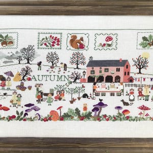 May include: A cross-stitch embroidery design depicting a charming autumn scene with a pink farmhouse, trees, and people gathered around a table. The word "AUTUMN" is stitched in the centre of the design.