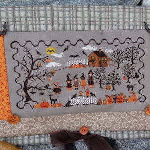 May include: A cross-stitch embroidery featuring a Halloween scene with a small house, a tree, and children dressed as witches. The embroidery is framed with a brown fabric border and has a scalloped edge. The background is a light gray fabric with a subtle pattern.