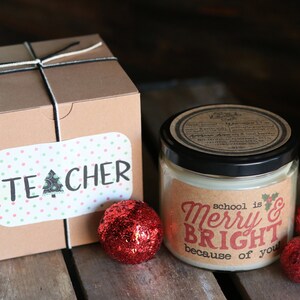 May include: A brown box wrapped with twine and a label that says "TEACHER". A candle in a glass jar with a brown label that says "school is Merry & Bright because of you". Three red glitter ornaments are on a wooden surface.