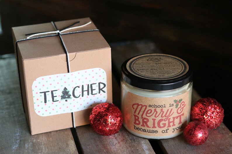 May include: A brown box wrapped with twine and a label that says "TEACHER". A candle in a glass jar with a brown label that says "school is Merry & Bright because of you". Three red glitter ornaments are on a wooden surface.